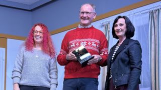 Ein Mann in einem roten Pullover hält eine Auszeichnung in den Händen, während er zwischen zwei lächelnden Frauen steht. Sie scheinen alle in einem Raum mit einer weißen Wand zu sein.