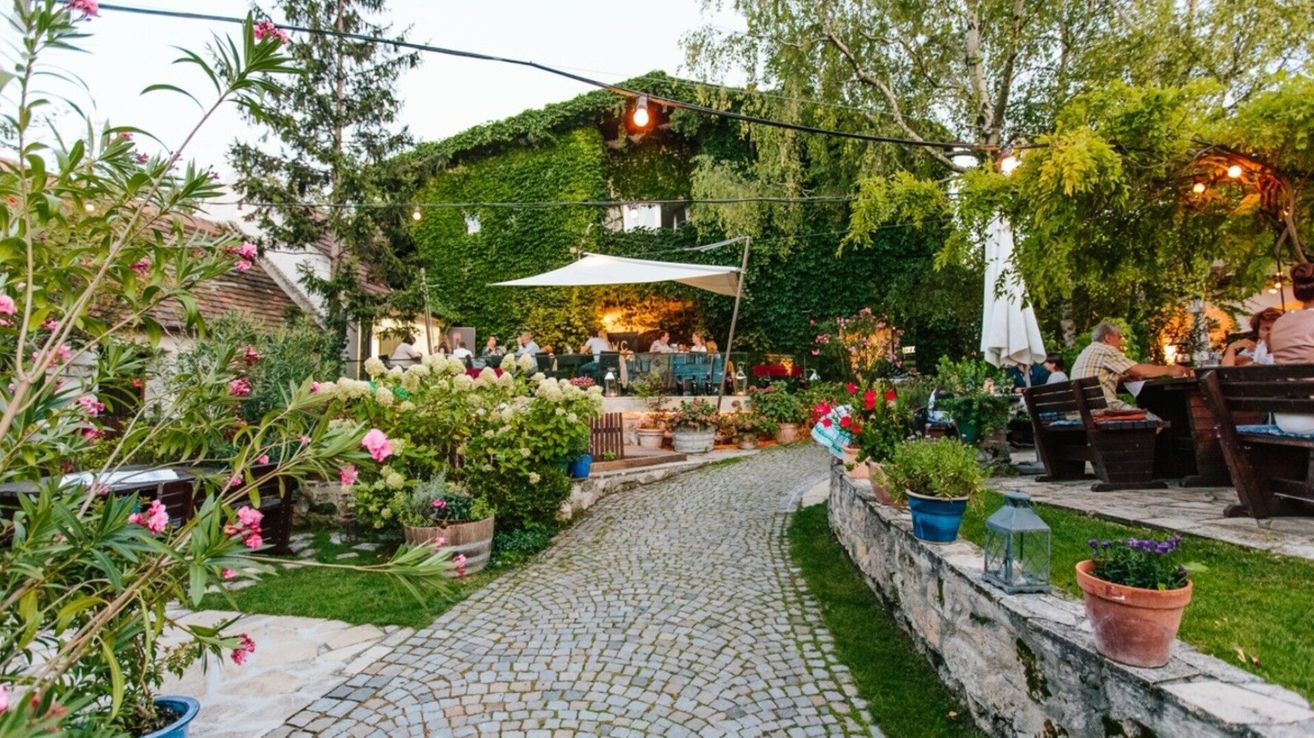Ein Außenrestaurant mit einem Kopfsteinpflasterweg, umgeben von üppiger Grünfläche, Blumen und Efeu-bewachsenen Mauern. Menschen sitzen an Tischen unter einem weißen Baldachin.