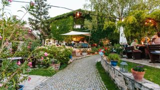 Ein Außenrestaurant mit einem Kopfsteinpflasterweg, umgeben von üppiger Grünfläche, Blumen und Efeu-bewachsenen Mauern. Menschen sitzen an Tischen unter einem weißen Baldachin.