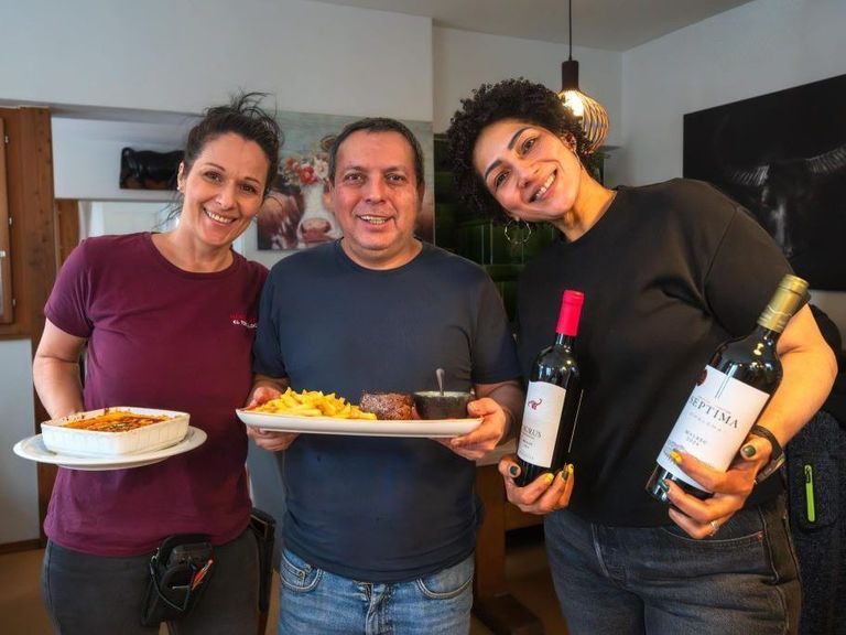 Drei lächelnde Personen posieren in einem Esszimmer. Ein Mann hält einen Teller mit Steak, Pommes und einem Löffel. Zwei Frauen halten Weinflaschen, eine mit grünem Nagellack.