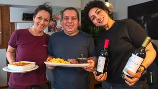 Drei lächelnde Personen posieren in einem Esszimmer. Ein Mann hält einen Teller mit Steak, Pommes und einem Löffel. Zwei Frauen halten Weinflaschen, eine mit grünem Nagellack.