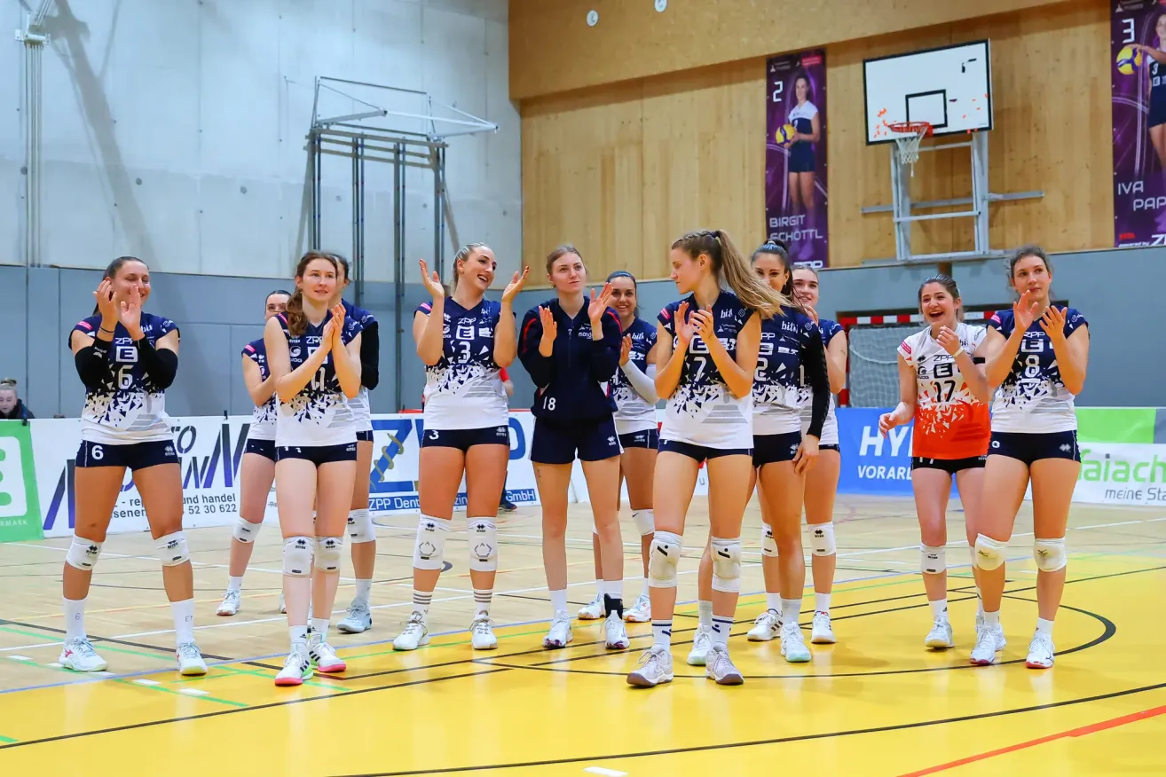 Eine Gruppe von weiblichen Volleyballspielerinnen steht in einer Reihe und klatscht in die Hände. Sie tragen passende Trikots mit Nummern auf dem Rücken. Der Platz hat einen gelben Boden und einen Basketballkorb im Hintergrund.