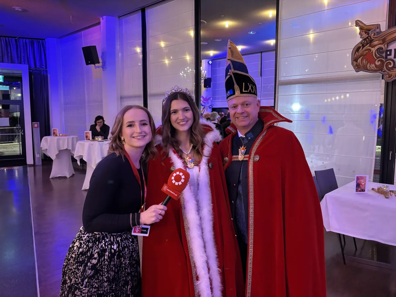 Drei Personen stehen in einem Raum mit Party-Dekoration. Eine Frau hält ein Mikrofon und interviewt eine Frau in einem roten Umhang und einen Mann in einem roten Umhang. Dahinter stehen Tische mit weißen Tischdecken.