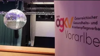 Eine Bühneneinrichtung mit Beleuchtungsequipment und einem weißen Vorhang. Ein rosafarbener Banner mit dem Text 'ogkv' und 'Gesundheits' ist sichtbar.
