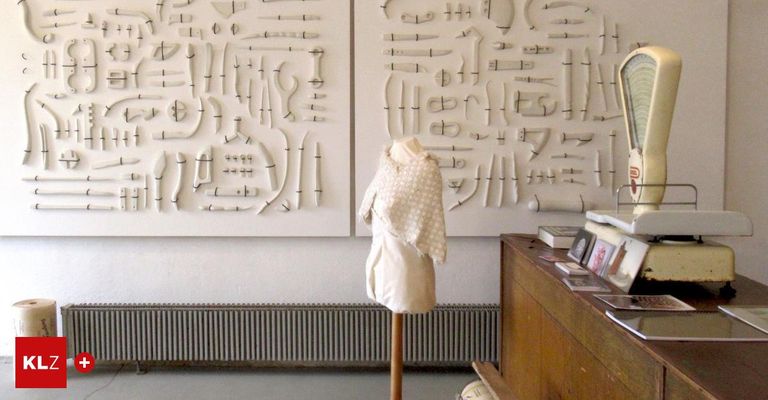 Ein weißes Kleidungsstück auf einem Mannequin steht vor zwei großen weißen Tafeln, die jeweils zahlreiche kleine, kunstvoll geschnitzte Objekte aufweisen. Rechts davon befindet sich ein hölzerner Schreibtisch mit Büchern und Bildern.