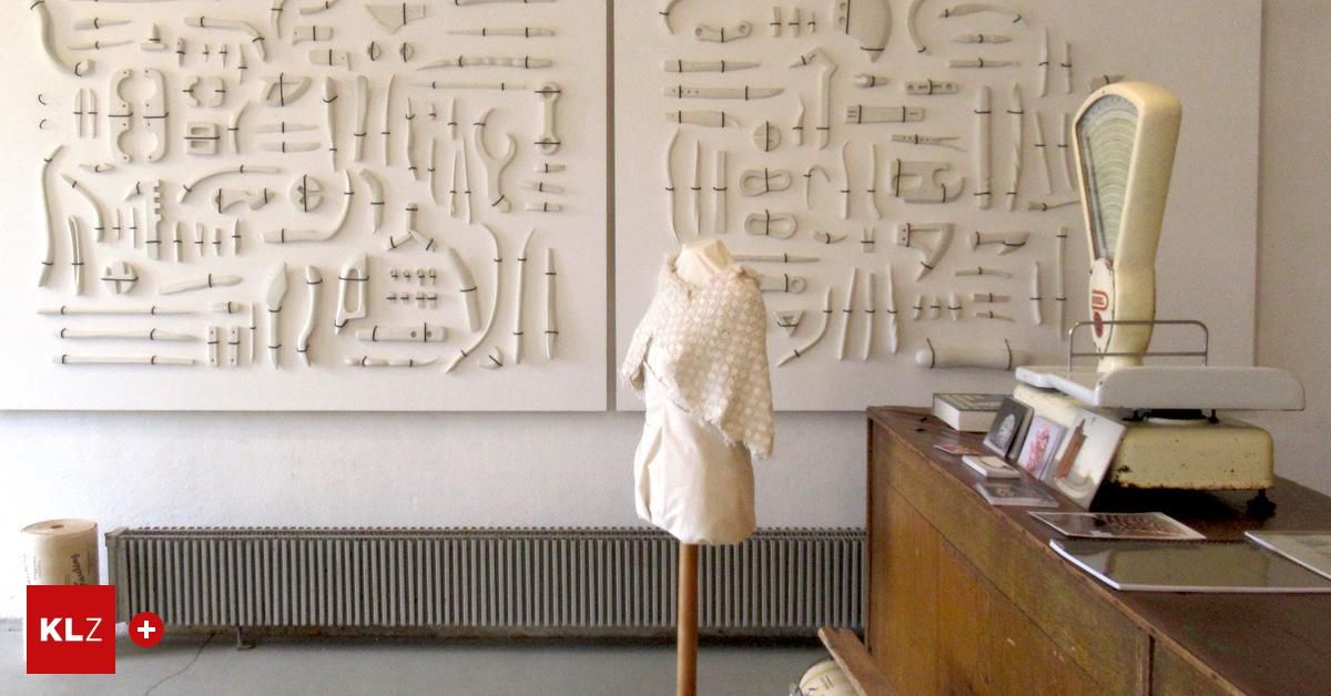 Ein weißes Kleidungsstück auf einem Mannequin steht vor zwei großen weißen Tafeln, die jeweils zahlreiche kleine, kunstvoll geschnitzte Objekte aufweisen. Rechts davon befindet sich ein hölzerner Schreibtisch mit Büchern und Bildern.