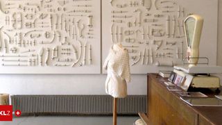 Ein weißes Kleidungsstück auf einem Mannequin steht vor zwei großen weißen Tafeln, die jeweils zahlreiche kleine, kunstvoll geschnitzte Objekte aufweisen. Rechts davon befindet sich ein hölzerner Schreibtisch mit Büchern und Bildern.