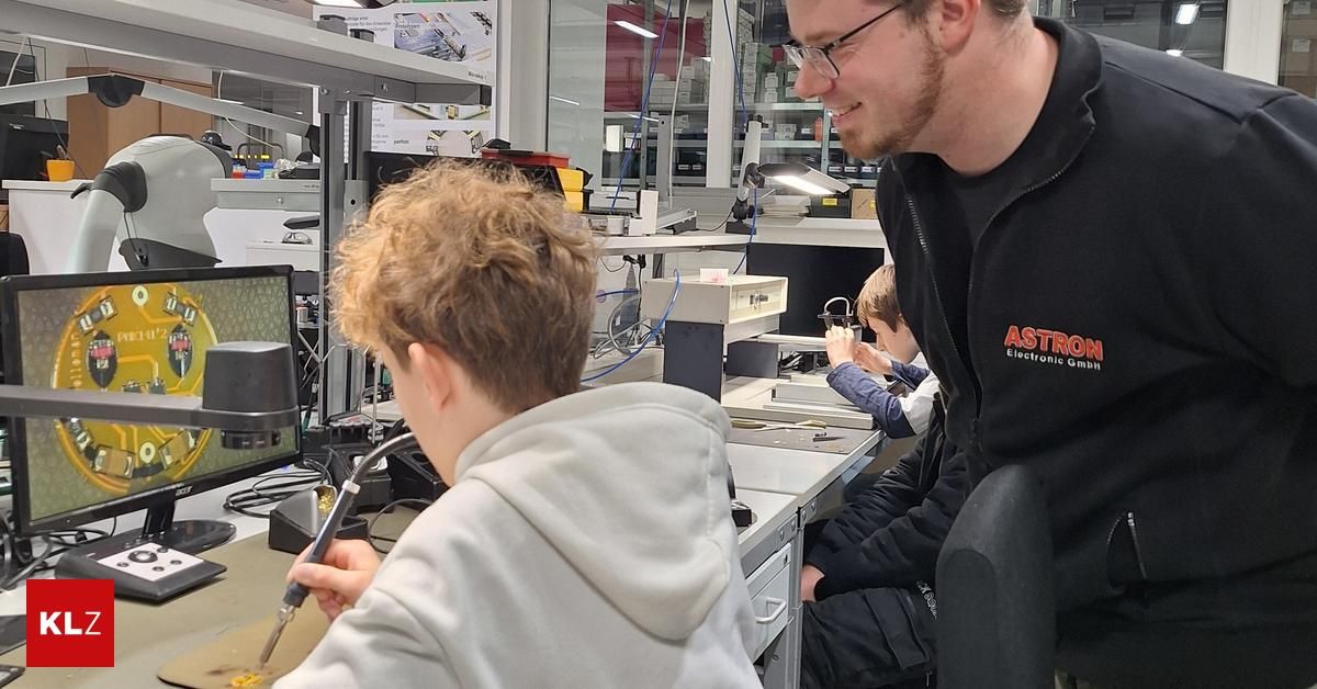 In einem Labor beobachtet ein Mann, wie ein Junge eine Schaltung lötet. Dahinter sitzt ein Kind, und Geräte reihen sich an den Regalen.