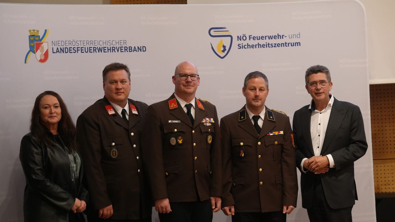 Vier Männer in Militäruniformen stehen vor einem weißen Banner mit einem Logo. Der Mann auf der linken Seite trägt eine Medaille und hat ein lächelndes Gesicht.