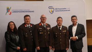 Vier Männer in Militäruniformen stehen vor einem weißen Banner mit einem Logo. Der Mann auf der linken Seite trägt eine Medaille und hat ein lächelndes Gesicht.