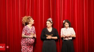 Drei Frauen stehen auf einer Bühne, halten Papiere und haben einen roten Vorhang im Hintergrund. Die Frau auf der linken Seite lächelt und hält ein Mikrofon. Die mittlere Frau hält ein Papier mit einem Mikrofon. Die Frau auf der rechten Seite sieht ernst aus und hält ein Papier.