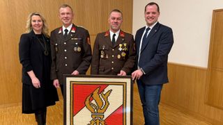 Drei Männer in Uniformen stehen zusammen und lächeln. Der Mann in der Mitte hält ein gerahmtes Emblem mit einem roten, weißen und gelben Design.