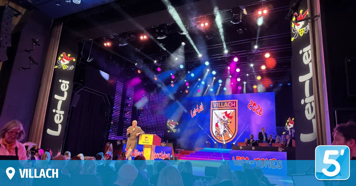 Ein Mann steht an einem Rednerpult auf der Bühne, mit einer großen Leinwand hinter ihm, auf der die Worte 'Villach 2026' und ein Logo stehen. Ein Publikum sitzt unten. Bühnenlichter in Rot, Blau und Lila beleuchten die Szene.