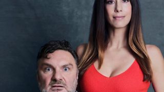 Ein Mann und eine Frau stehen eng beieinander und posieren für ein Foto. Der Mann schaut nach links, während die Frau lächelt und direkt in die Kamera schaut. Die Frau trägt ein rotes Tanktop.