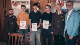 Vier Männer stehen vor einer Holzwand mit einem Banner und halten Urkunden in der Hand. Der Mann auf der rechten Seite trägt eine Militäruniform. Die beiden Männer in der Mitte tragen schwarze Hemden und Jeans. Der Mann auf der linken Seite trägt eine orangefarbene Kapuzenjacke.