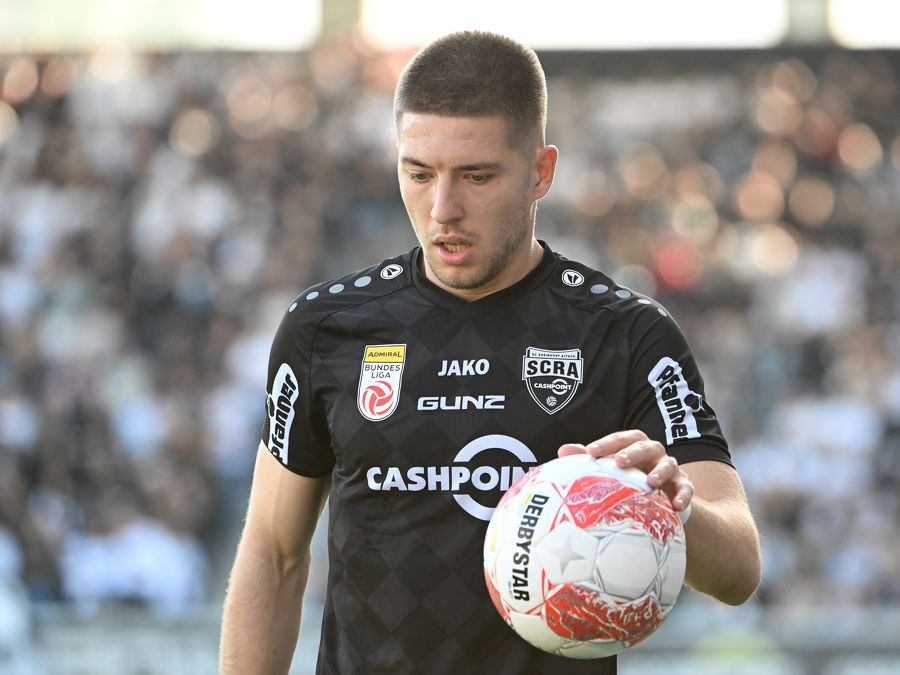 Ein Fußballspieler hält einen Fußball in der Hand und trägt ein schwarzes Trikot mit mehreren Sponsorenlogos.