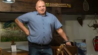 Ein Mann mit einem Schnurrbart und einer Uhr steht im Inneren, lehnt sich an ein hölzerne Handlauf und trägt ein blaues Hemd und Jeans. Hinter ihm befindet sich eine hölzerne Wand mit einem Holzschild.