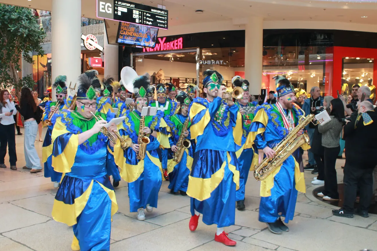 Eine Marschkapelle in blauen und gelben Kostümen spielt Musikinstrumente auf der Straße. Sie gehen vor einem Geschäft mit einem digitalen Schild.