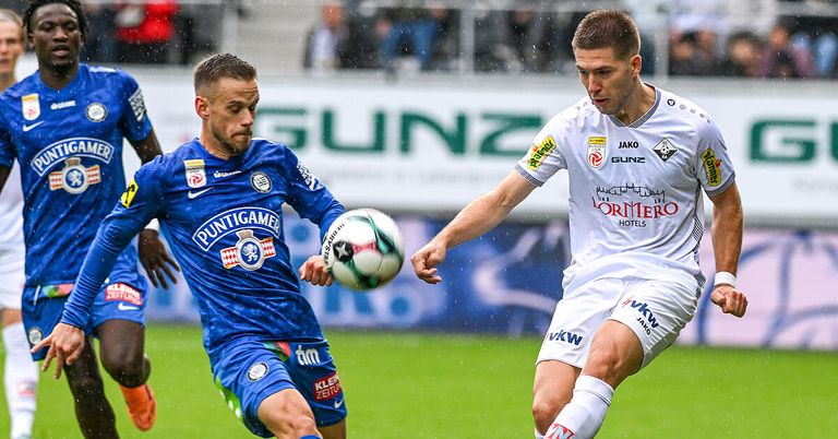 Zwei Fußballspieler sind auf einem Feld. Ein Spieler trägt ein blaues Trikot und der andere ein weißes. Sie jagen einem Ball im Regen nach.