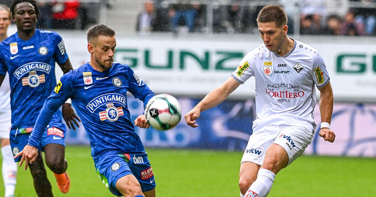 Zwei Fußballspieler sind auf einem Feld. Ein Spieler trägt ein blaues Trikot und der andere ein weißes. Sie jagen einem Ball im Regen nach.