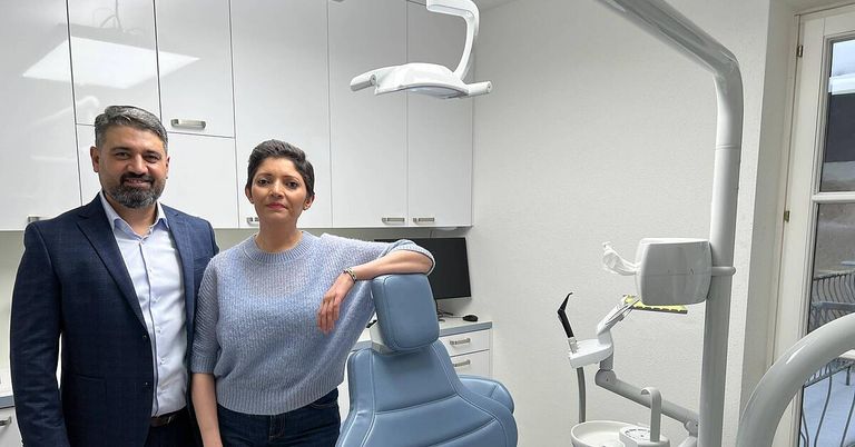 Ein Mann und eine Frau stehen in einem Zahnarztpraxis, wobei sich die Frau auf einen blauen Zahnarztstuhl stützt. Hinter ihnen befinden sich weiße Schränke und ein Monitor auf einem Schreibtisch.