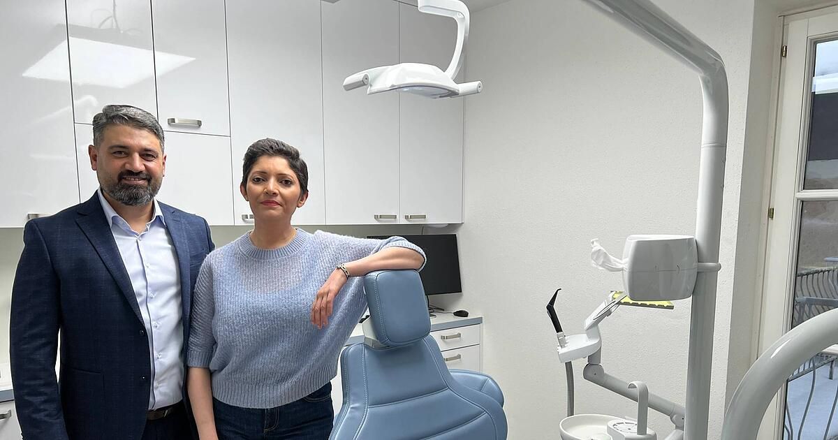 Ein Mann und eine Frau stehen in einem Zahnarztpraxis, wobei sich die Frau auf einen blauen Zahnarztstuhl stützt. Hinter ihnen befinden sich weiße Schränke und ein Monitor auf einem Schreibtisch.