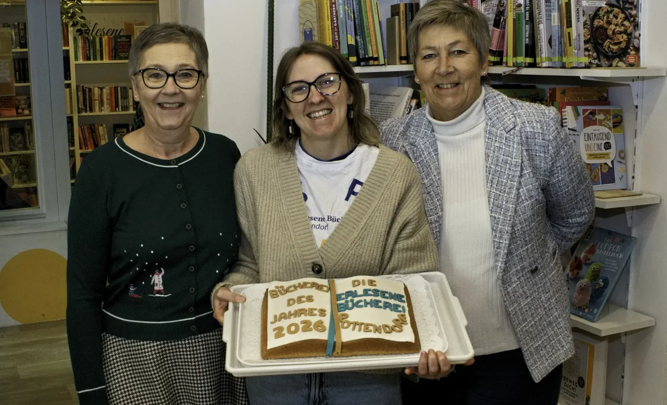 Drei Frauen stehen zusammen und halten einen Kuchen, auf dem 'Bucherei des Jahres 2026' steht. Die Frau in der Mitte trägt eine Brille und einen Pullover. Die anderen beiden Frauen tragen Rollkragenpullover und Blazer. Dahinter steht ein Bücherregal voller Bücher.