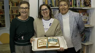 Drei Frauen stehen zusammen und halten einen Kuchen, auf dem 'Bucherei des Jahres 2026' steht. Die Frau in der Mitte trägt eine Brille und einen Pullover. Die anderen beiden Frauen tragen Rollkragenpullover und Blazer. Dahinter steht ein Bücherregal voller Bücher.
