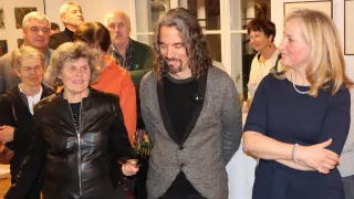 Mehrere Menschen stehen in einem Raum, wahrscheinlich bei einer Veranstaltung. Ein Mann mit langen Haaren und einem Schnurrbart lächelt und schaut nach vorne. Hinter ihm lächelt eine Frau und schaut nach links. Andere Menschen sind im Hintergrund.