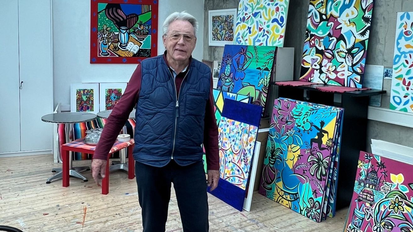 Ein älterer Mann mit Brille steht in einem Kunstatelier. Er trägt eine blaue Weste und schwarze Hose. Hinter ihm hängen bunte Gemälde an der Wand.
