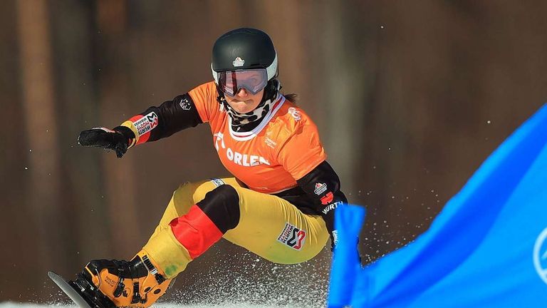 Ein Snowboarder in einem orange-schwarzen Outfit mit 'Orlen' und 'Wurth', trägt einen schwarzen Helm und eine Brille, ist in der Luft und es spritzt Schnee um ihn herum.