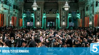 Eine große Menschenmenge in formeller Kleidung tanzt in einem großen Saal mit grüner Beleuchtung und Kronleuchtern. Die Menge scheint zu feiern.