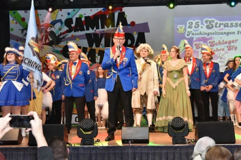 Eine Gruppe von Menschen in Kostümen steht auf der Bühne mit Scheinwerfern und einem Banner, das 'Samma Wilda' steht. Der Mann in der Mitte hält ein Mikrofon.