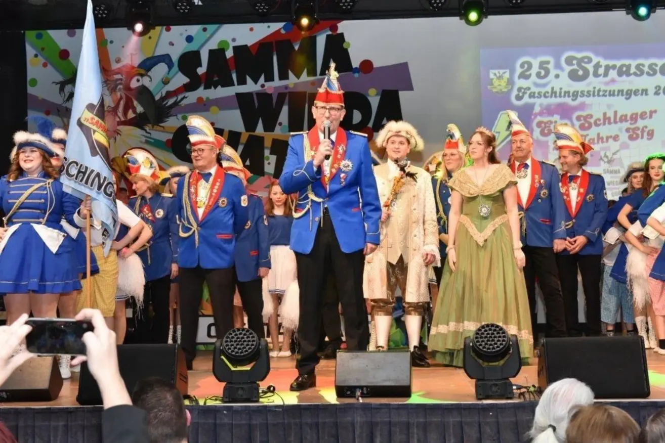 Eine Gruppe von Menschen in Kostümen steht auf der Bühne mit Scheinwerfern und einem Banner, das 'Samma Wilda' steht. Der Mann in der Mitte hält ein Mikrofon.