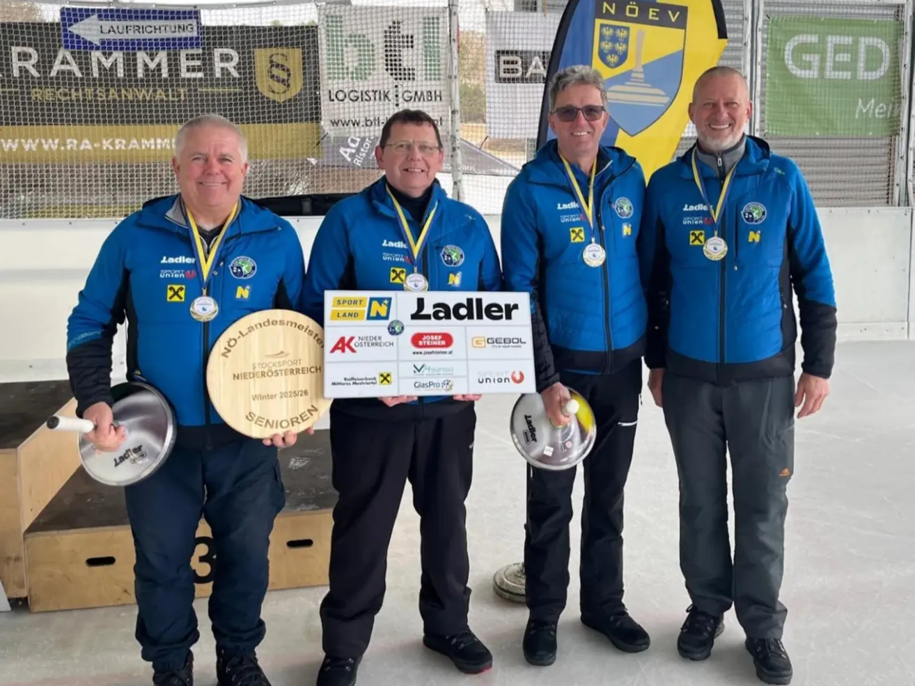 Vier Männer in blauen Jacken halten Medaillen und Trophäen. Sie stehen in einer Reihe, lächeln und posieren für ein Foto. Hinter ihnen befindet sich ein Banner mit dem Text "NOEV".