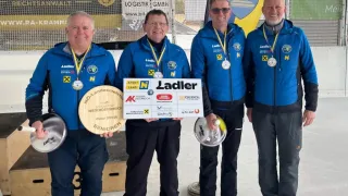 Vier Männer in blauen Jacken halten Medaillen und Trophäen. Sie stehen in einer Reihe, lächeln und posieren für ein Foto. Hinter ihnen befindet sich ein Banner mit dem Text "NOEV".