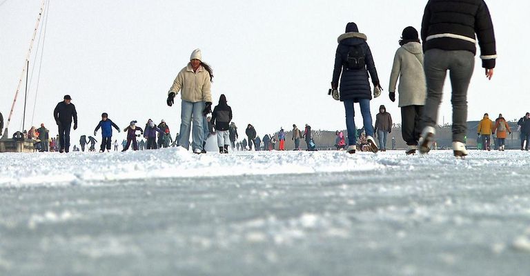 Viele Menschen gehen auf einer verschneiten Oberfläche, einige skaten. Sie sind warm gekleidet, mit Mützen, Handschuhen und Wintermänteln. Der Himmel ist klar.