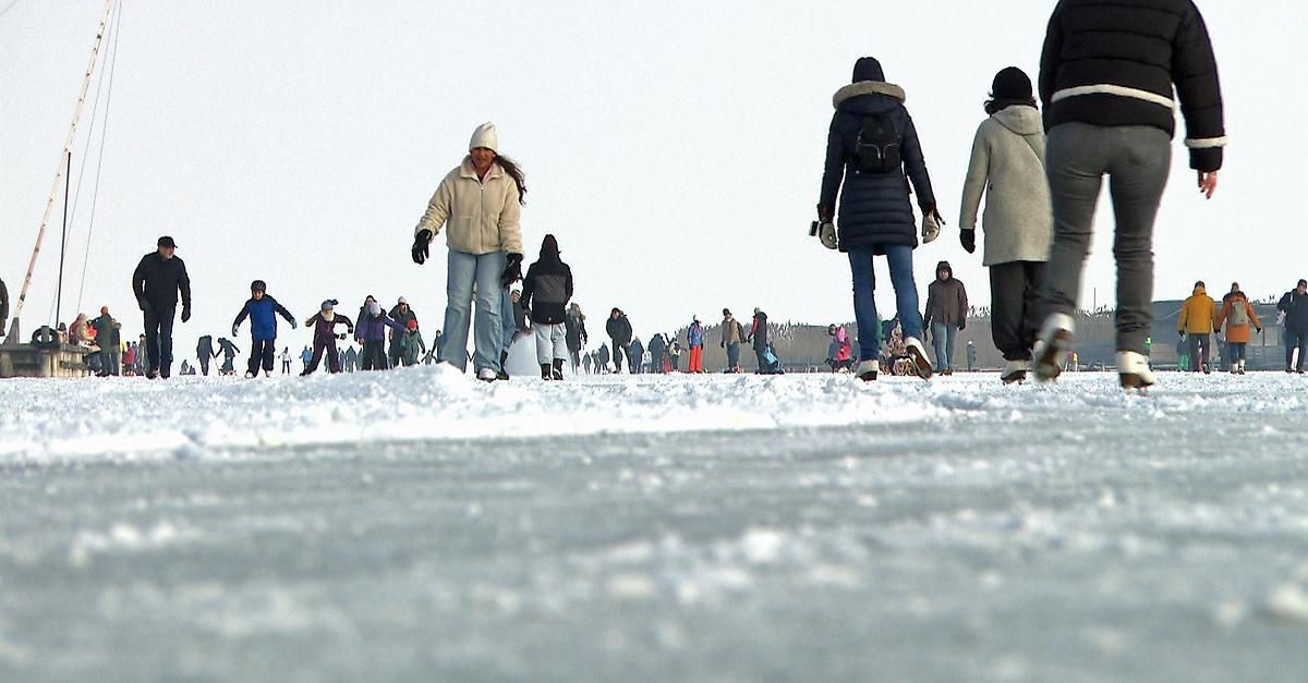 Viele Menschen gehen auf einer verschneiten Oberfläche, einige skaten. Sie sind warm gekleidet, mit Mützen, Handschuhen und Wintermänteln. Der Himmel ist klar.