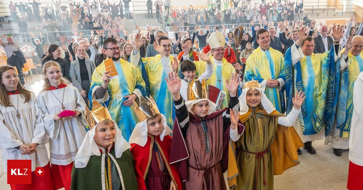 Eine Gruppe von Kindern in festlichen Kostümen und Kronen winkt zur Kamera. Dahinter winken Priester und Männer in Roben ebenfalls. Eine Menge im Hintergrund schaut zu.