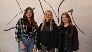 Drei junge Frauen stehen nebeneinander vor einer weißen Wand mit abstrakten schwarzen Mustern. Die linke trägt ein kariertes Hemd und Jeans, die mittlere eine schwarze Kapuzenjacke mit weißem Text und die rechte eine Jeansjacke und eine Halskette.