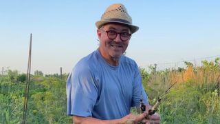 Ein Mann mit Strohhut und Brille hält eine Pflanze. Er lächelt und scheint sich in einem Grasfeld zu befinden.