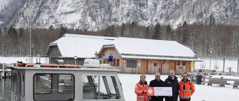 Vier Männer in orange Weste stehen vor einem Gebäude und einem Boot, einer hält einen Scheck. Dahinter bildet ein verschneiter Berg die Kulisse.