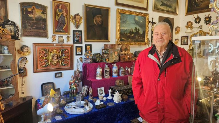 Ein älterer Mann steht in einem Raum voller verschiedener Gegenstände und Kunstwerke und lächelt. Hinter ihm hängen mehrere gerahmte Gemälde, darunter eines von einem Mann mit Hut. Darunter befinden sich zahlreiche Gegenstände auf einem blauen Tisch, wie Figuren, Schachteln und Glasobjekte.