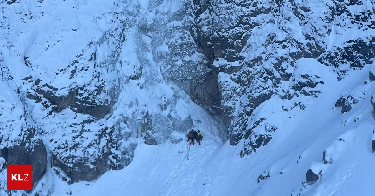 Zwei Personen erklimmen einen verschneiten Berg und benutzen Seile und Kletterausrüstung. Sie tragen Helme und Rucksäcke.