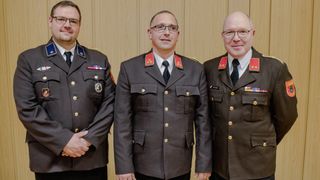 Drei Männer in Militäruniformen stehen zusammen. Der Mann in der Mitte trägt eine Brille und einen Ring. Die Männer an den Seiten tragen Medaillen.