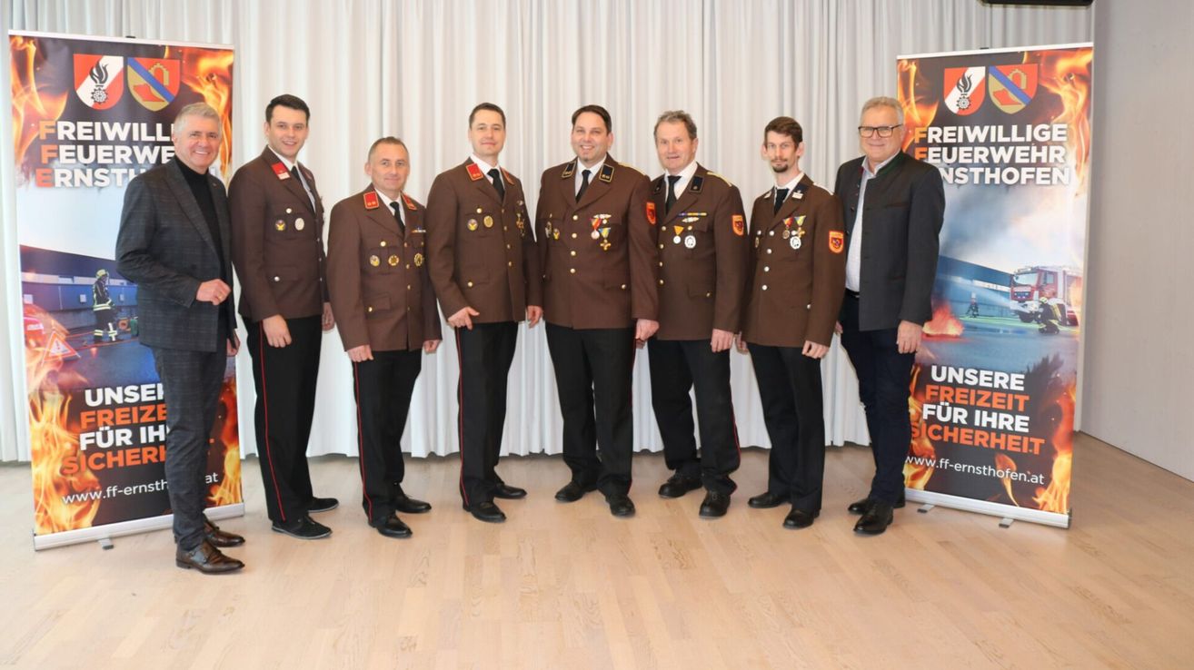 Sieben Männer in Militäruniformen stehen in einer Reihe vor einem weißen Vorhang. Sie lächeln und sehen in die Kamera. Sie tragen Medaillen und Abzeichen an ihren Uniformen.