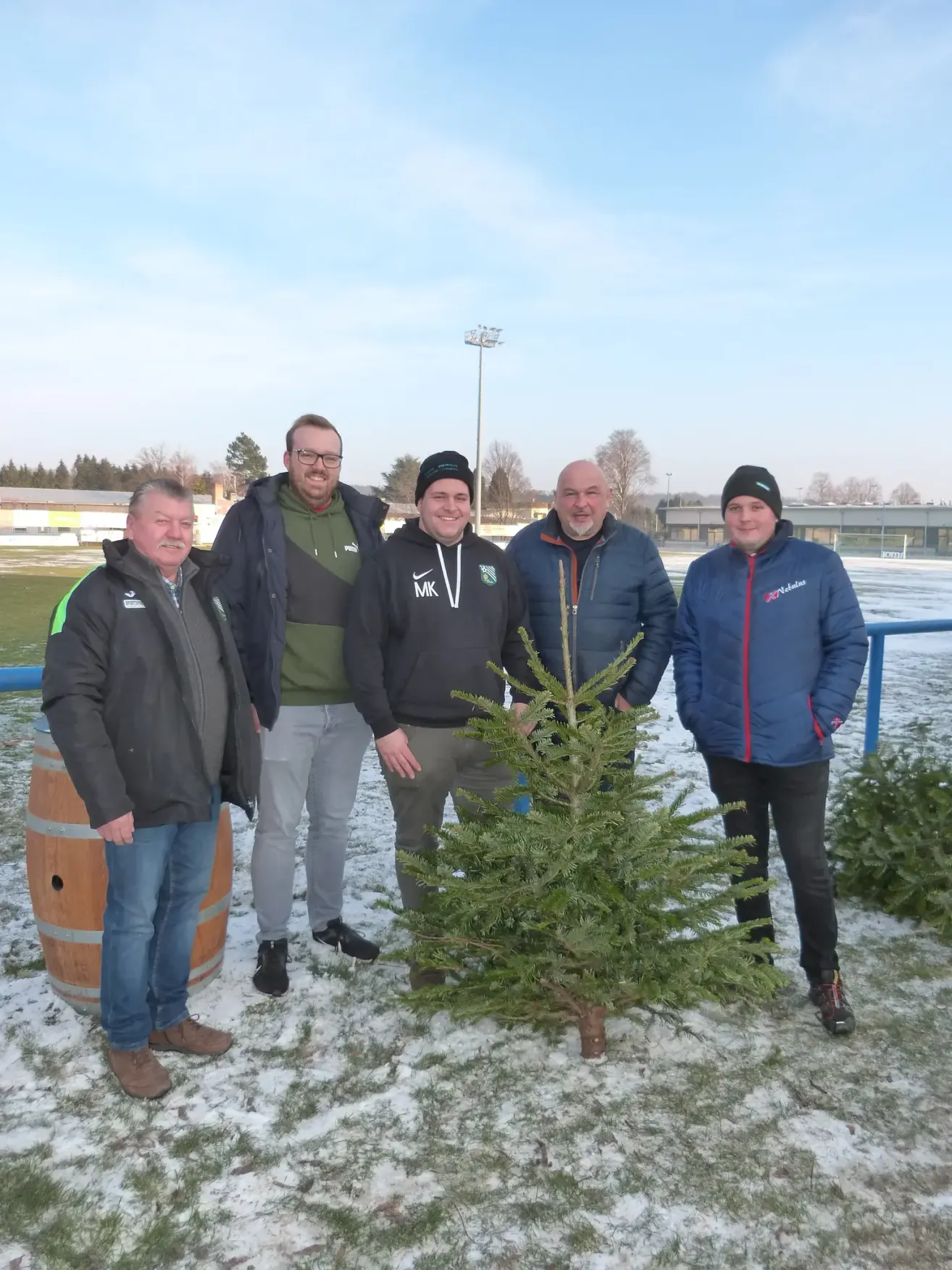 Fünf Männer stehen in Winterkleidung, lächelnd für ein Foto auf einem verschneiten Feld, mit einem kleinen Weihnachtsbaum in der Mitte und einem Fass hinter dem linken Mann.
