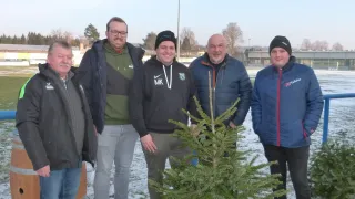 Fünf Männer stehen in Winterkleidung, lächelnd für ein Foto auf einem verschneiten Feld, mit einem kleinen Weihnachtsbaum in der Mitte und einem Fass hinter dem linken Mann.
