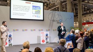 Ein Mann steht auf einer Bühne und hält eine Präsentation für ein Publikum, mit einer großen Leinwand dahinter. Die Leinwand zeigt eine reisebezogene Präsentation.
