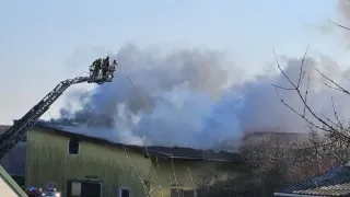 Feuerwehrleute stehen auf einer Leiter, die Wasser auf ein brennendes Gebäude sprüht. Rauch steigt vom Dach auf, und Menschen stehen am Boden.